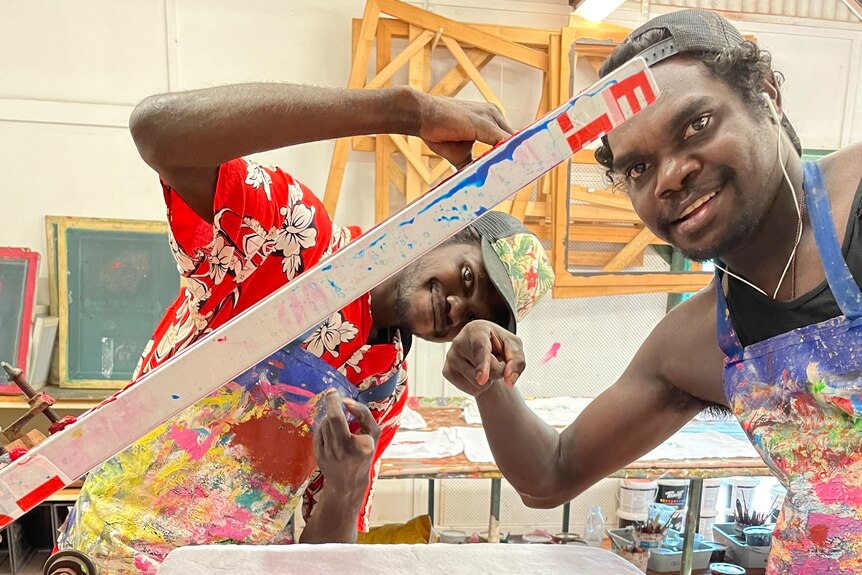 Two Aboriginal men smile and point at the camera while operating a screen printing machine