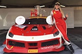 Female race car driver in overalls with her Ferrari race car and numerous trophies.