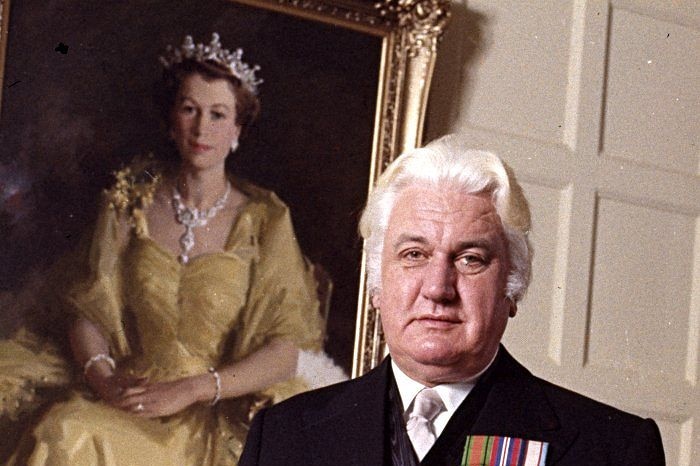 A man dressed in a suit with white hair standing in front of a painting of Queen Elizabeth II.