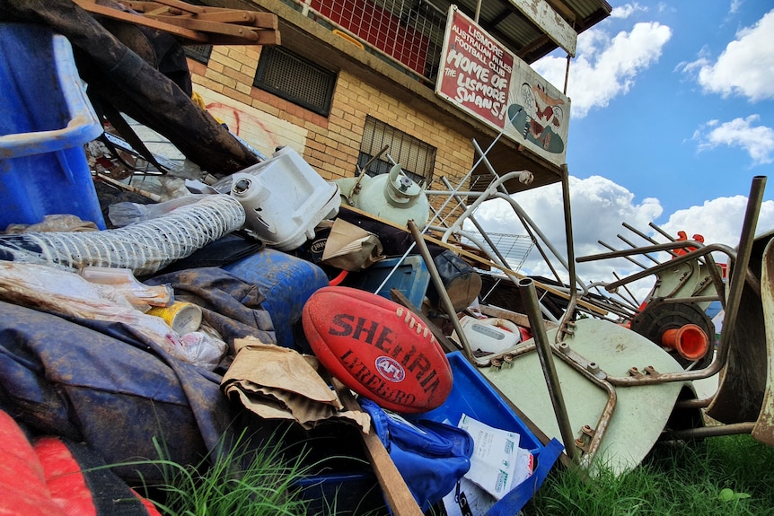 A red Sherrin football sits on a pile of rubbish. A building with Lismore Swans club signage is in the background.