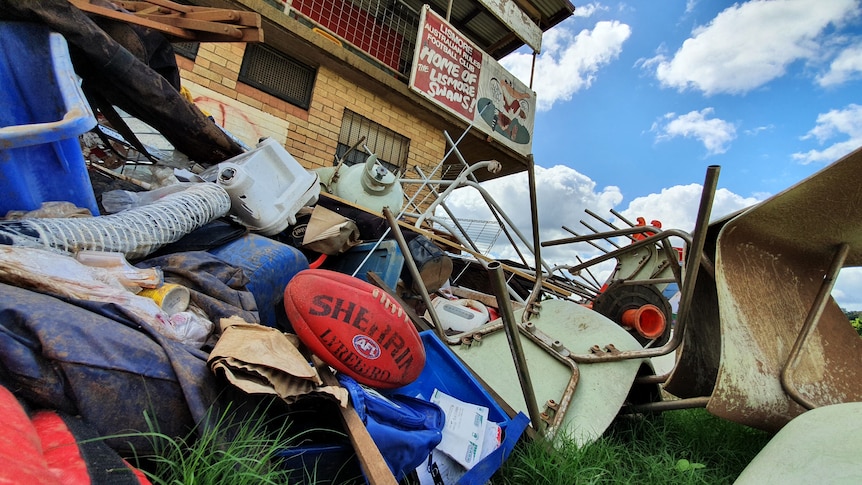 A red Sherrin football sits on a pile of rubbish. A building with Lismore Swans club signage is in the background.