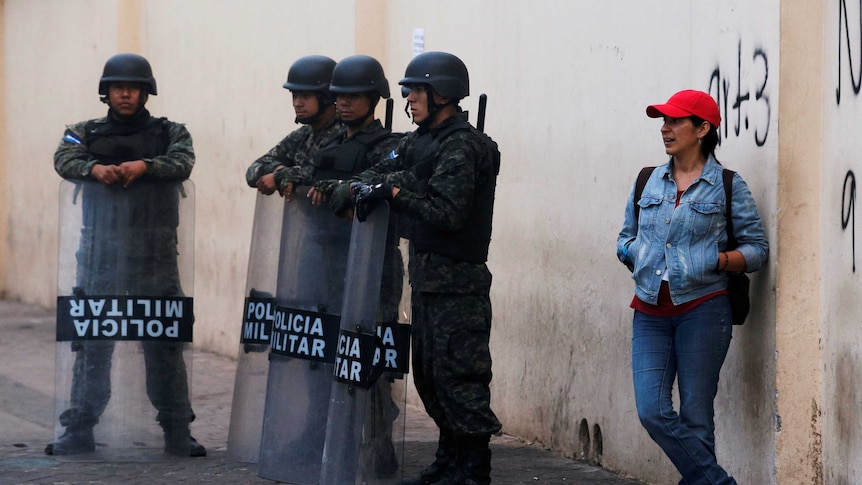 A woman leans against a wall next to armoured military police.