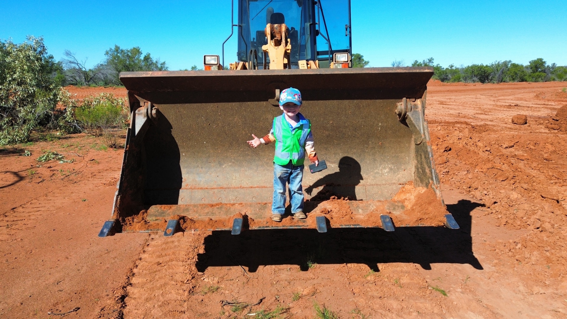 A young boy in jeans and a high vis jacket standing in the scoop of a bulldozer on a patch of dirt