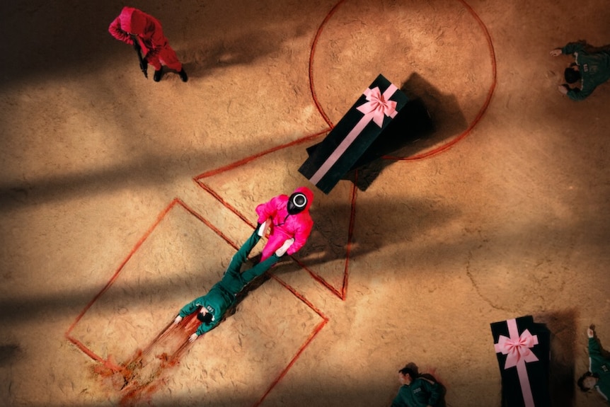 A person in a pink jumpsuit and black mask drags a body with blood next to a coffin