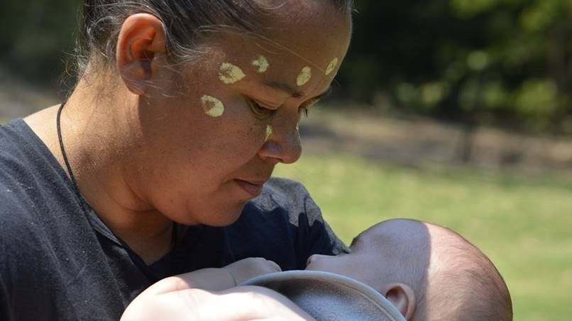 Close up of Melanie Briggs nursing a new born, she is holding it close and Melanie has traditional ochre painting on her face.