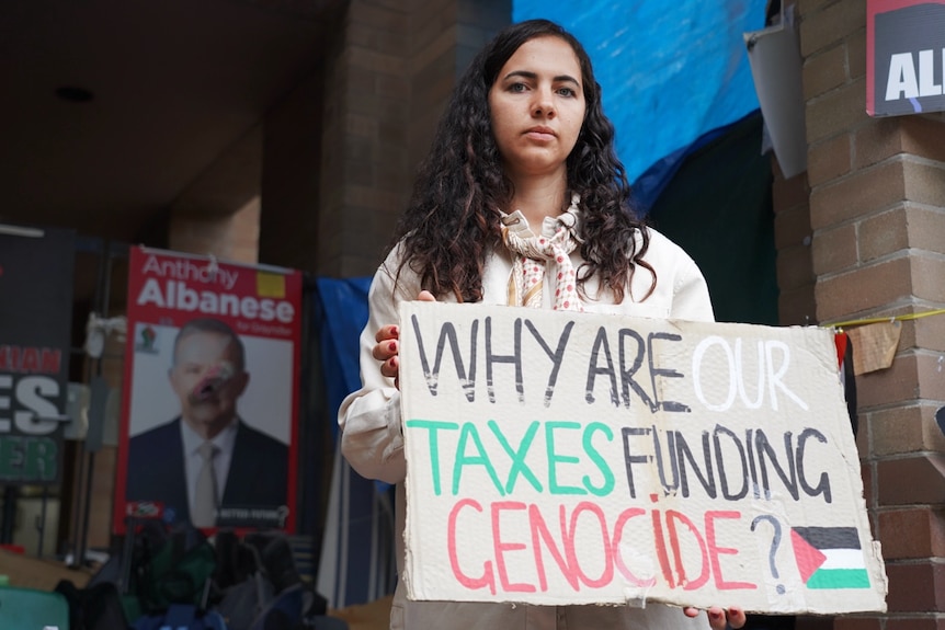 A woman stands with a sign that says 'Why are our taxes funding genocide'