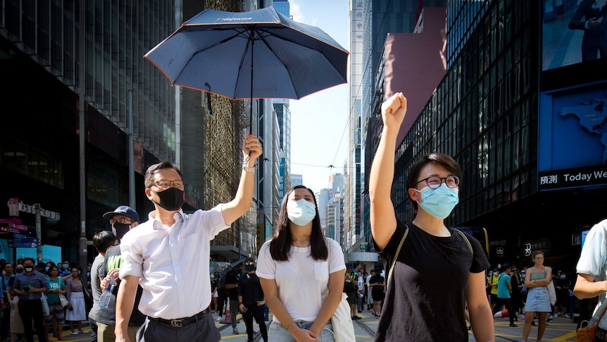 Three protesters stand close to the camera with masks on, one holding an umbrella.