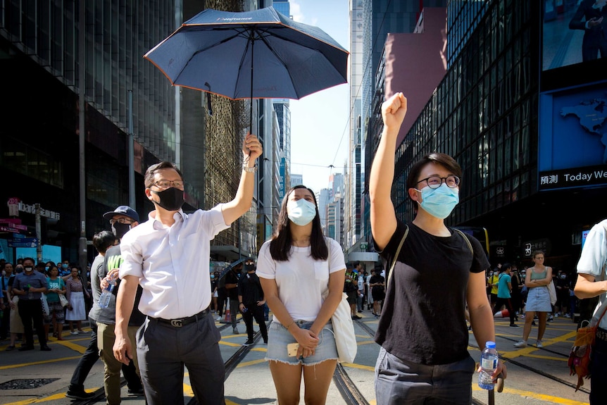 Three protesters stand close to the camera with masks on, one holding an umbrella.