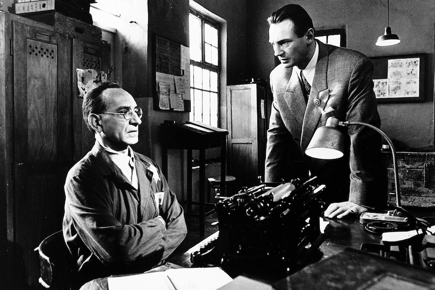 Two men in suits talk at a desk