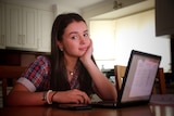 A teenage girl sitting at her laptop in her school uniform at the kitchen table. 