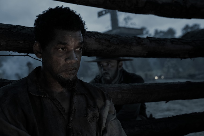 A desaturated image of an upset-looking Black man. A white man in a hat is visible behind a fence behind him.