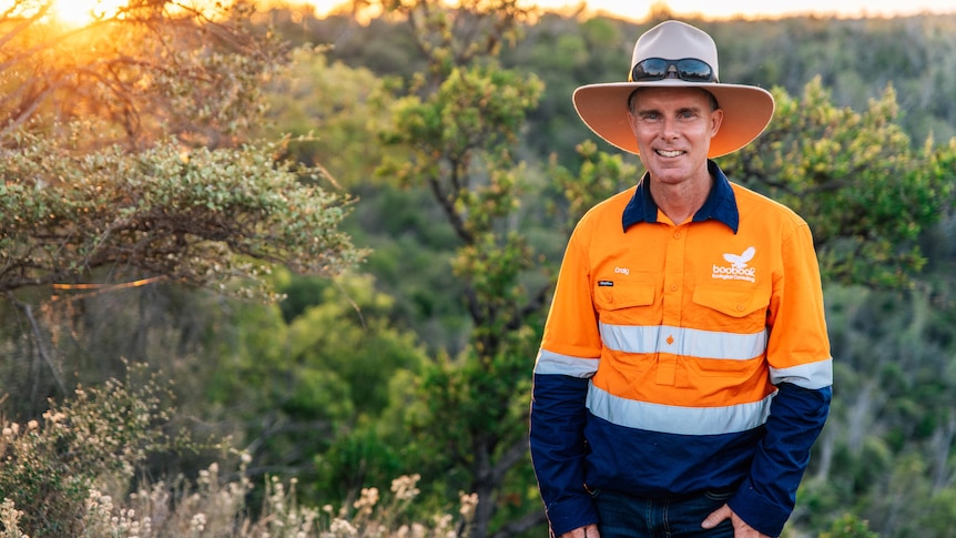 Man with a hat on and orange safety coloured shirt standing in the bush with the sun behind him.