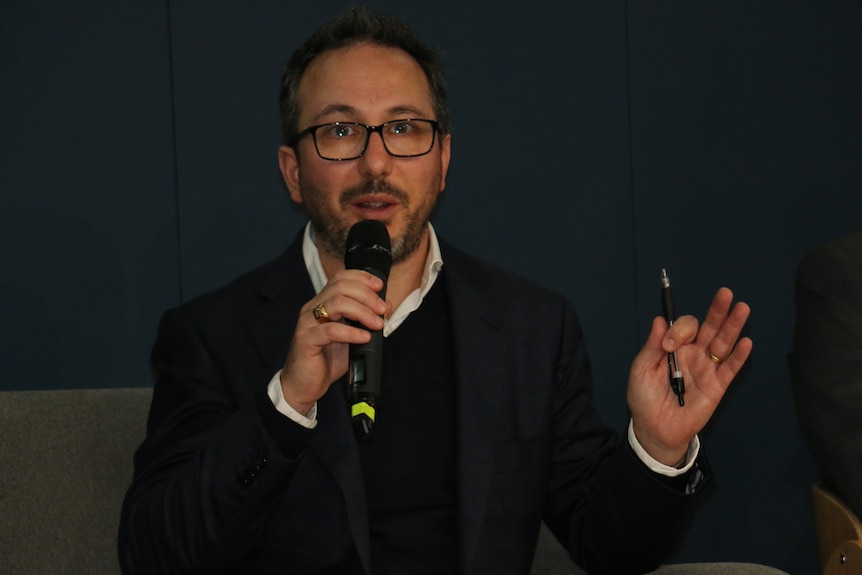 Lee wearing a dark jumper, holding a microphone in a room while talking.