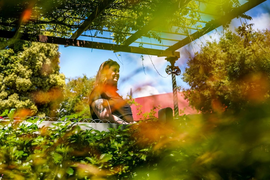 A woman rests against a walkway fence surrounded by lush green trees on a sunny day.