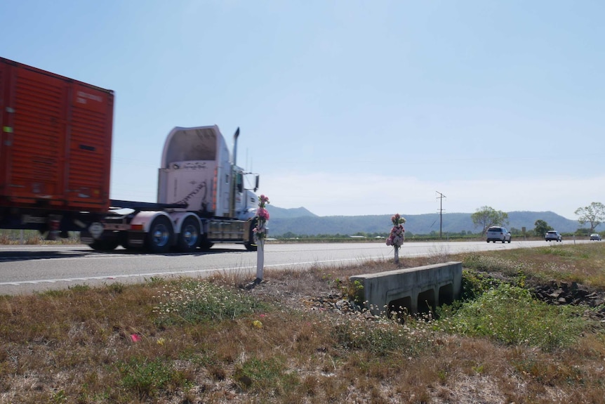 One truck and four cars driving away from the camera on a stretch of road behind two posts which are wrapped with fake flowers.