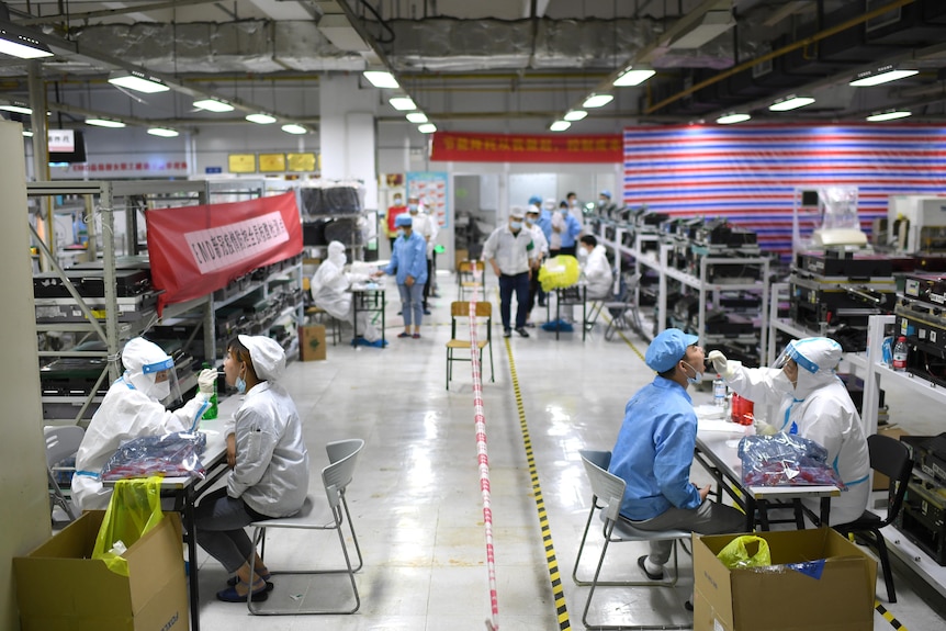 Two factory workers sit at separate desks, each across from a medical worker in PPE inserting a swab into their mouth