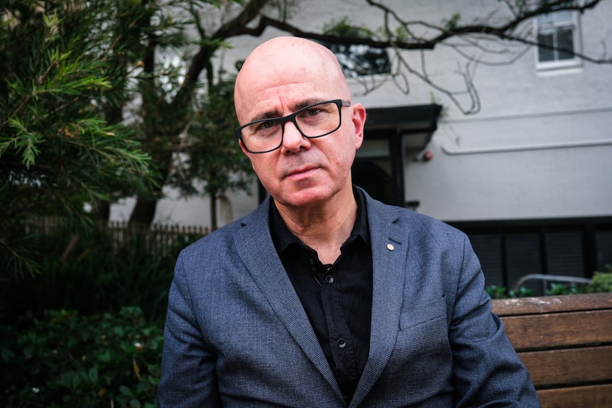 A bald, bespectacled man in a dark blazer stands on a street, looking concerned.