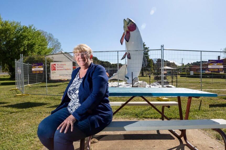An older woman sitting in front of a statue of a big trout which is under repair