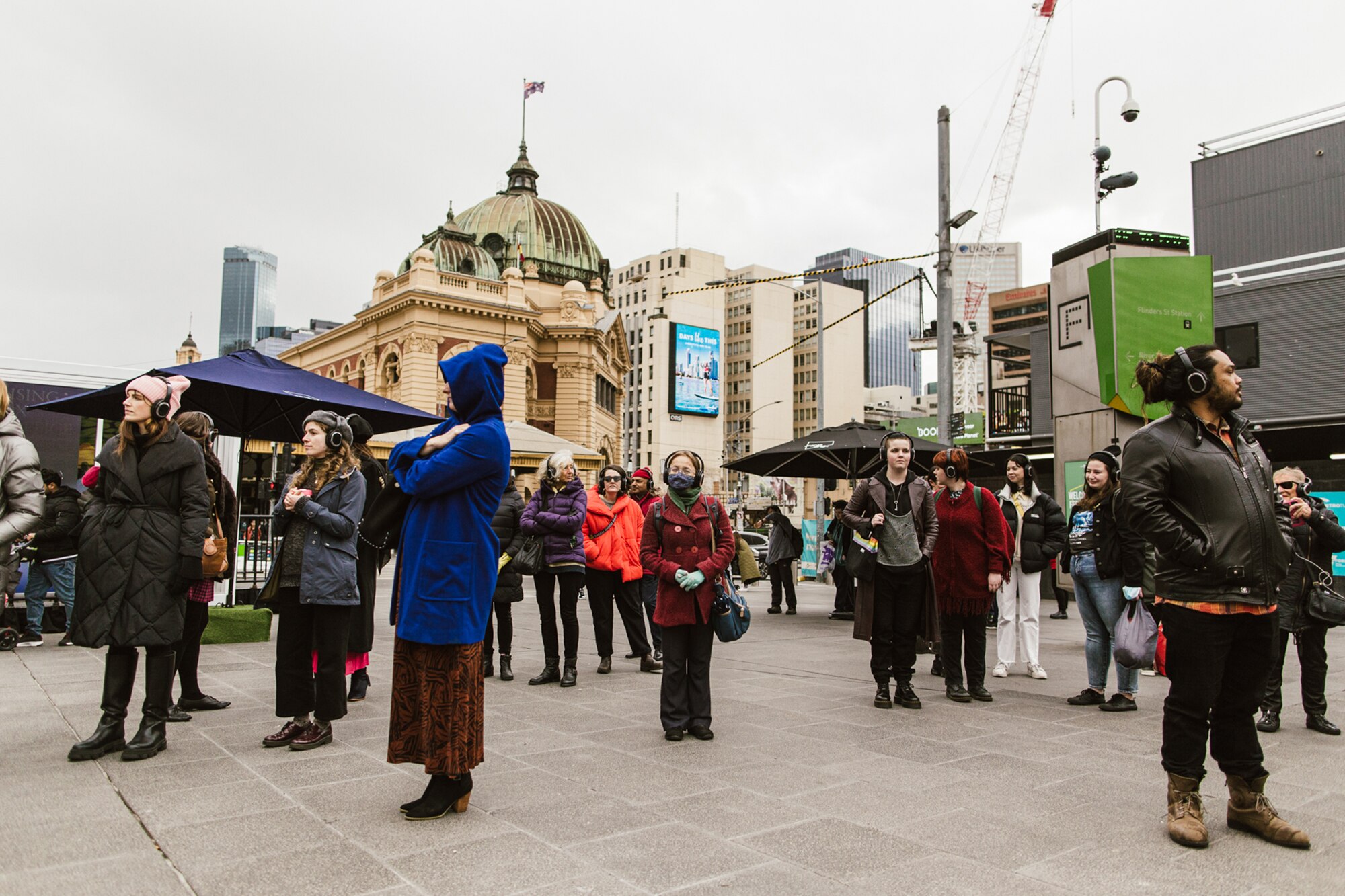 A crowd of people stand in Melbourne's Federation Square on a grey, overcast day. They are wearing headphones.