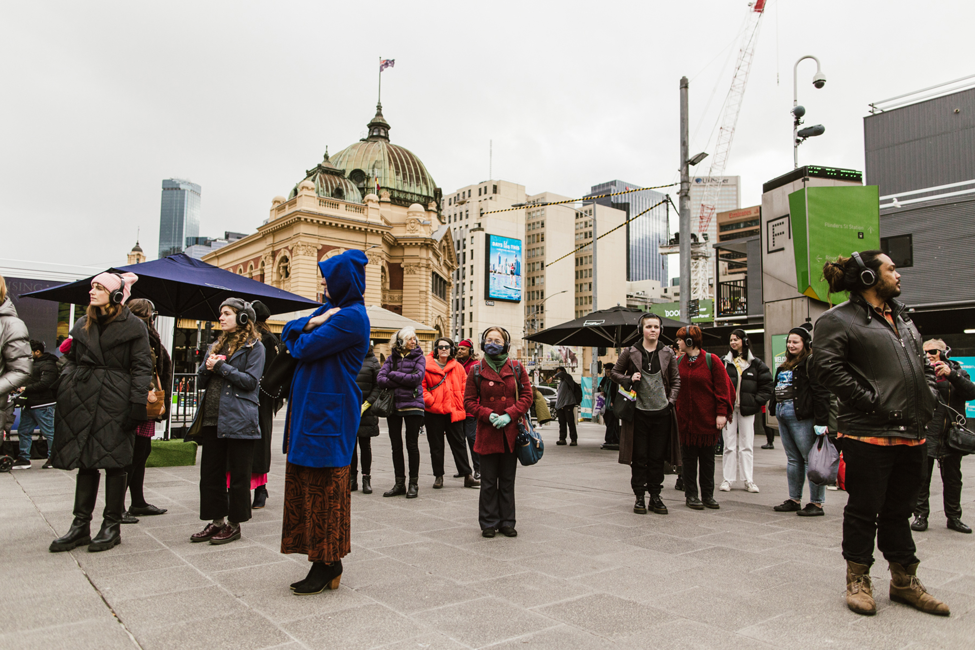 A crowd of people stand in Melbourne's Federation Square on a gray, overcast day.  They are wearing headphones.