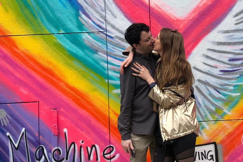 A man and woman embrace in front of a wall with a rainbow heart painted on