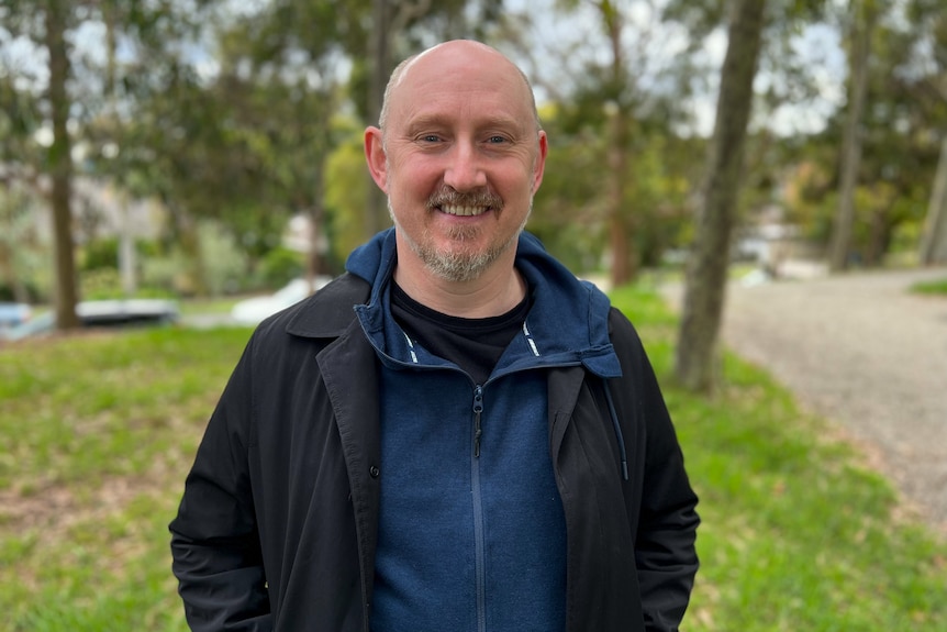 Dean stands in a park wearing a navy jumper and dark overcoat and smiles at the camera