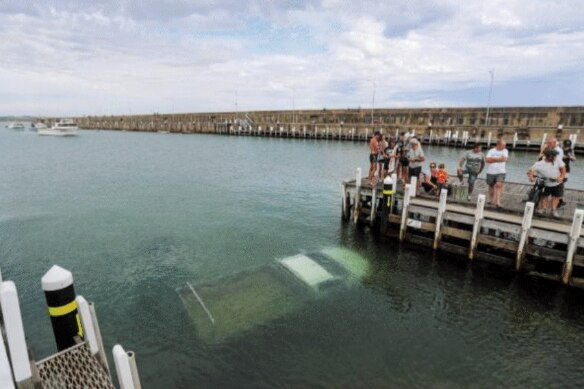 A group of poeple stand at a boat ramp, a large object that looks like a ute submerged in the water.