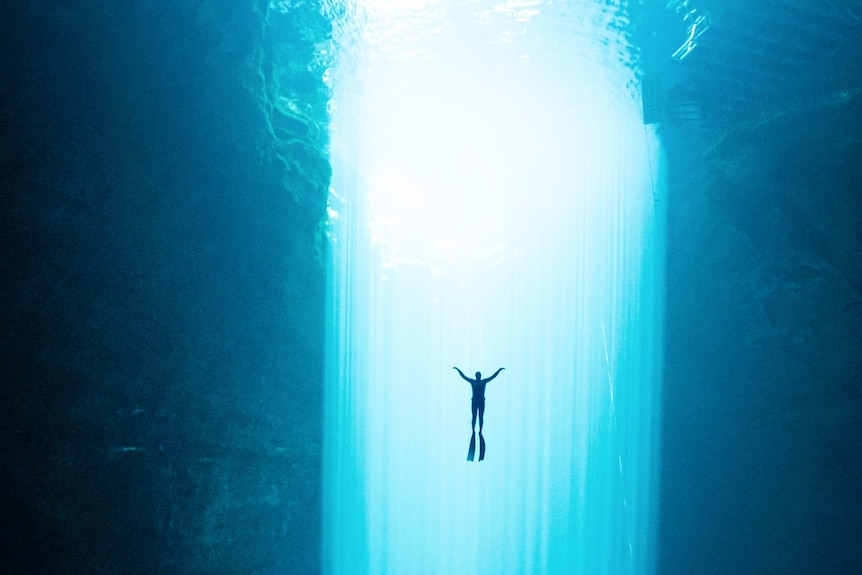 A diver in a wetsuit float towards the surface of a large light beam in turquoise water.