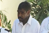 An Indigenous man wearing a white shirt walking between two security guards, wearing handcuffs.