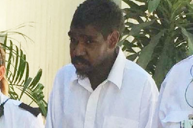 An Indigenous man wearing a white shirt walking between two security guards, wearing handcuffs.