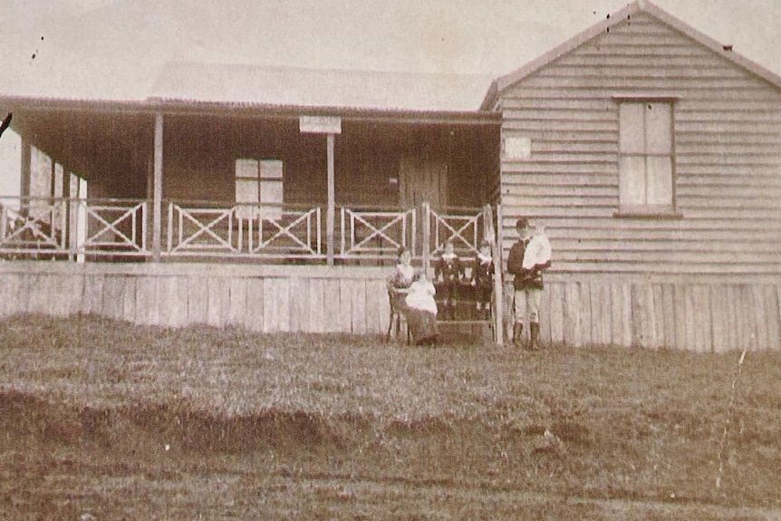 A black and white image showing an older timber building with an outside verandah and a family in 1900s style clothing.
