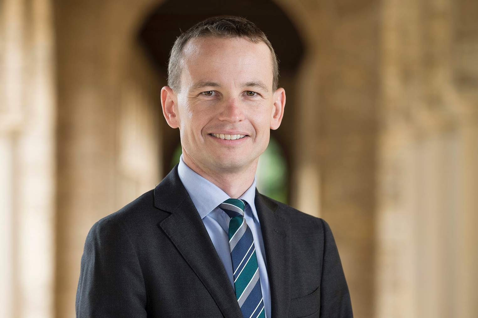 A head and shoulders shot of a man smiling wearing a dark suit, blue shirt and blue and green striped tie.