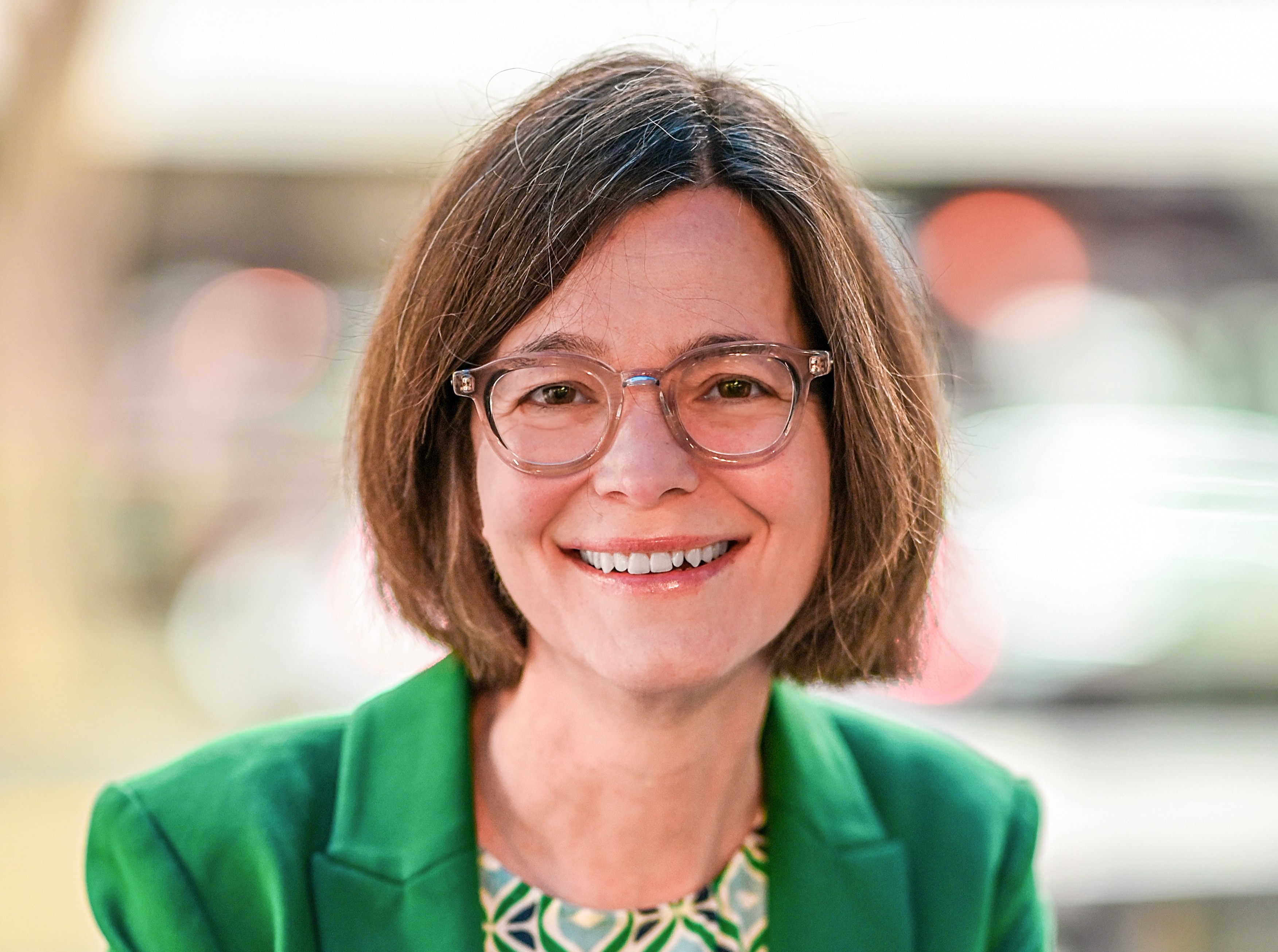 A headshot of a women with brown bobbed hair, glasses and green jacket.