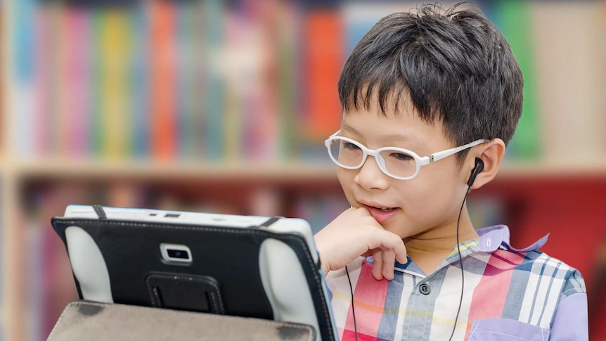 A young Asian boy with glasses and earphones watches something on a tablet that is propped up on a table.
