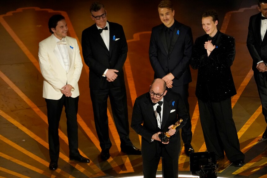 A white man in a suit speaks on a stage into a microphone while holding an Academy award. Five other men stand on the stage.