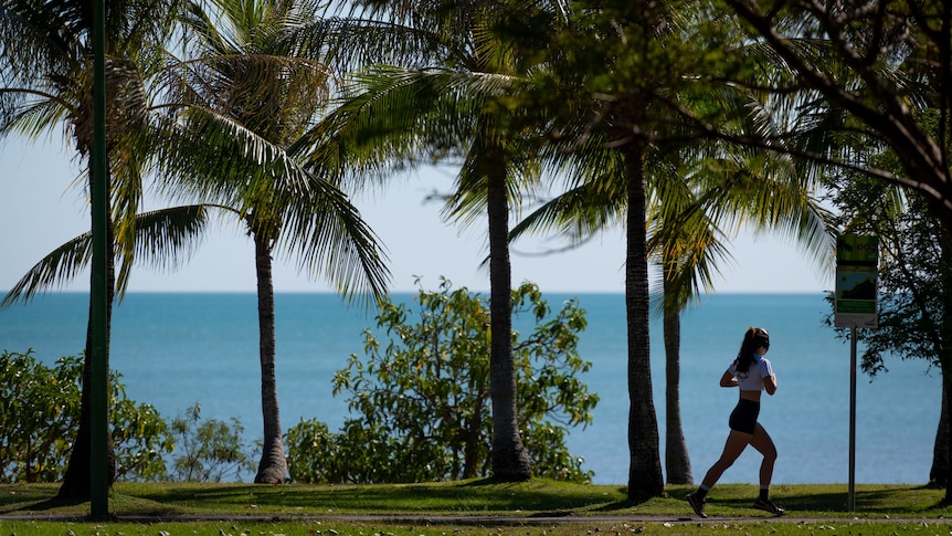 A photo of a young woman running on a path near a beach. There are palm trees and grass.