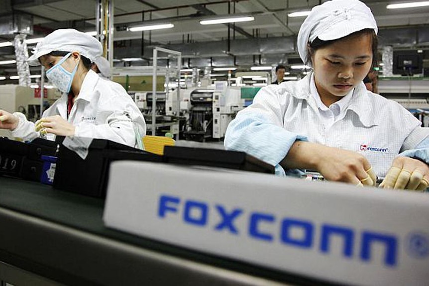 Two women in white outfits and yellow gloves work inside a factory, standing behind a sign that reads Foxconn 