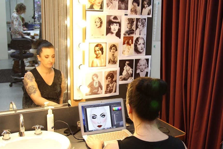 Woman sits before mirror looking at laptop, with photos pinned on wall