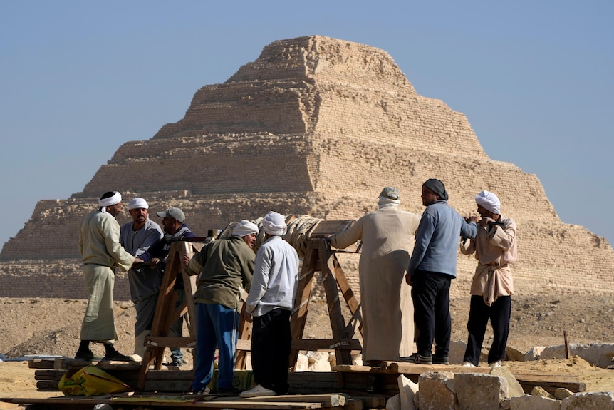 A group of men stand with a wooden structure as a pyramid looms in the background. 