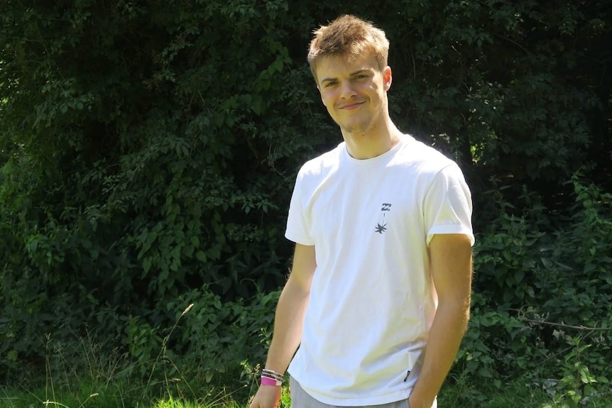 A young man in a white shirt, smiling in front of some trees.