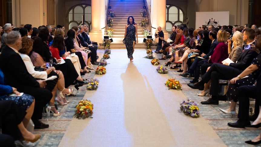 Shannon McGuire models a dress in front of a catwalk crowd in a big hall with pillars and a large staircase