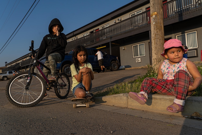 Two small children sit on the kerb outside a motel. Another sits on her bicycle behind them.