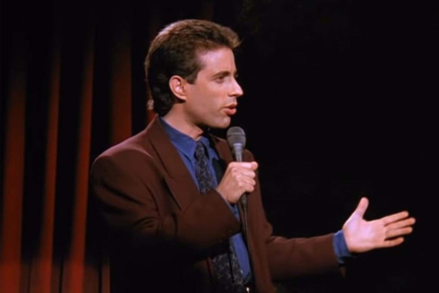 Jerry Seinfeld stands on stage in front of a red curtain, holding a microphone.