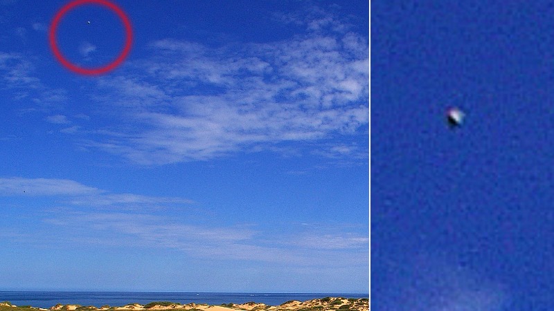 A small round object sits high in the sky above a beach with sand dunes in the foreground.
