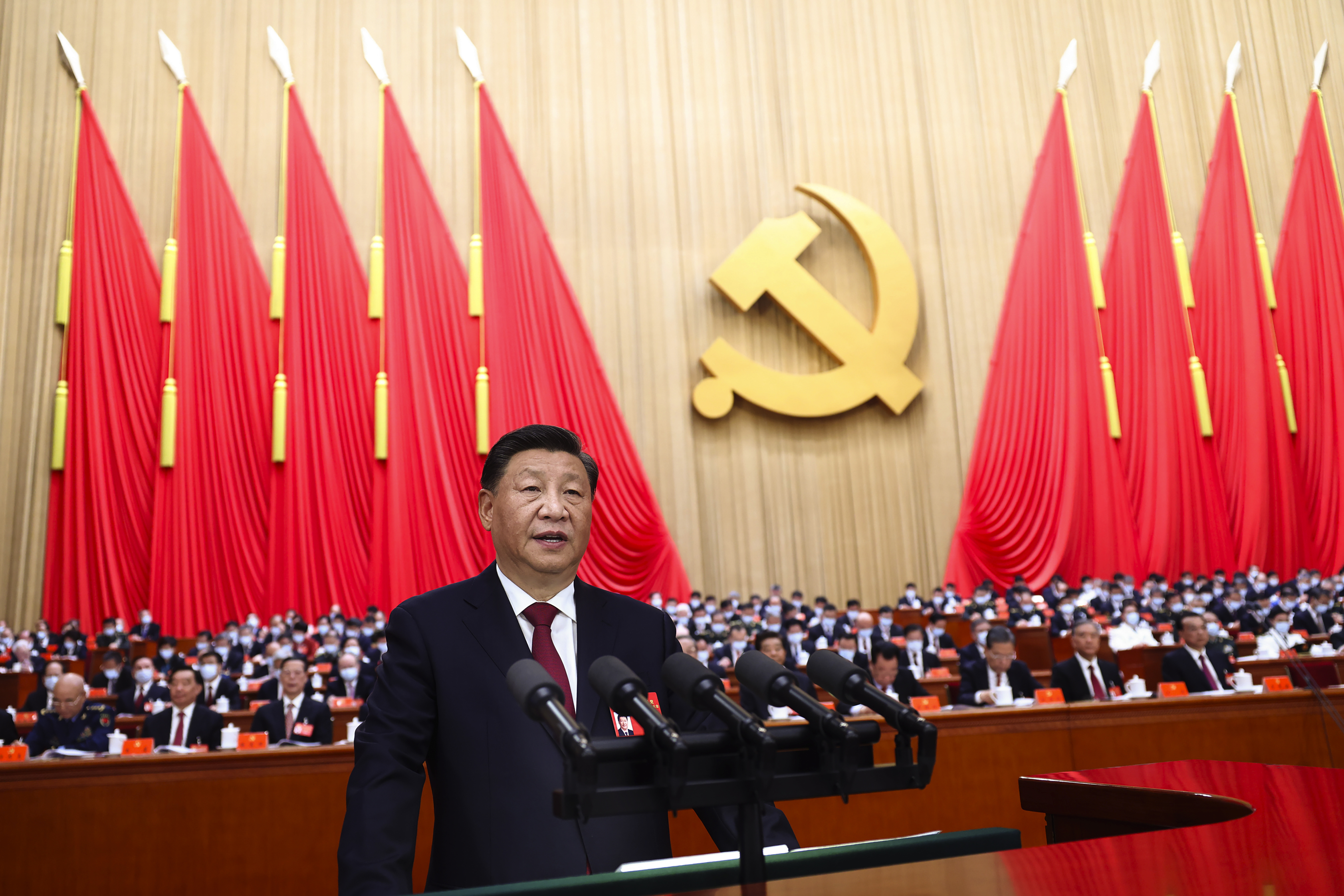 一个穿着深色西装的男人在一大群坐着的人群和共产主义服饰面前对着麦克风讲话。