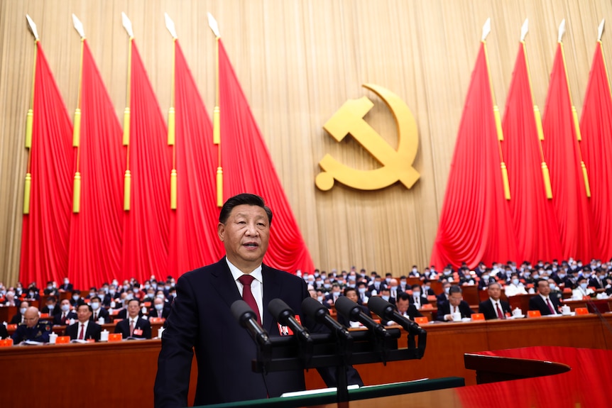 A man in a dark suit speaks into a mic in front of a large seated crowd and communist regalia.