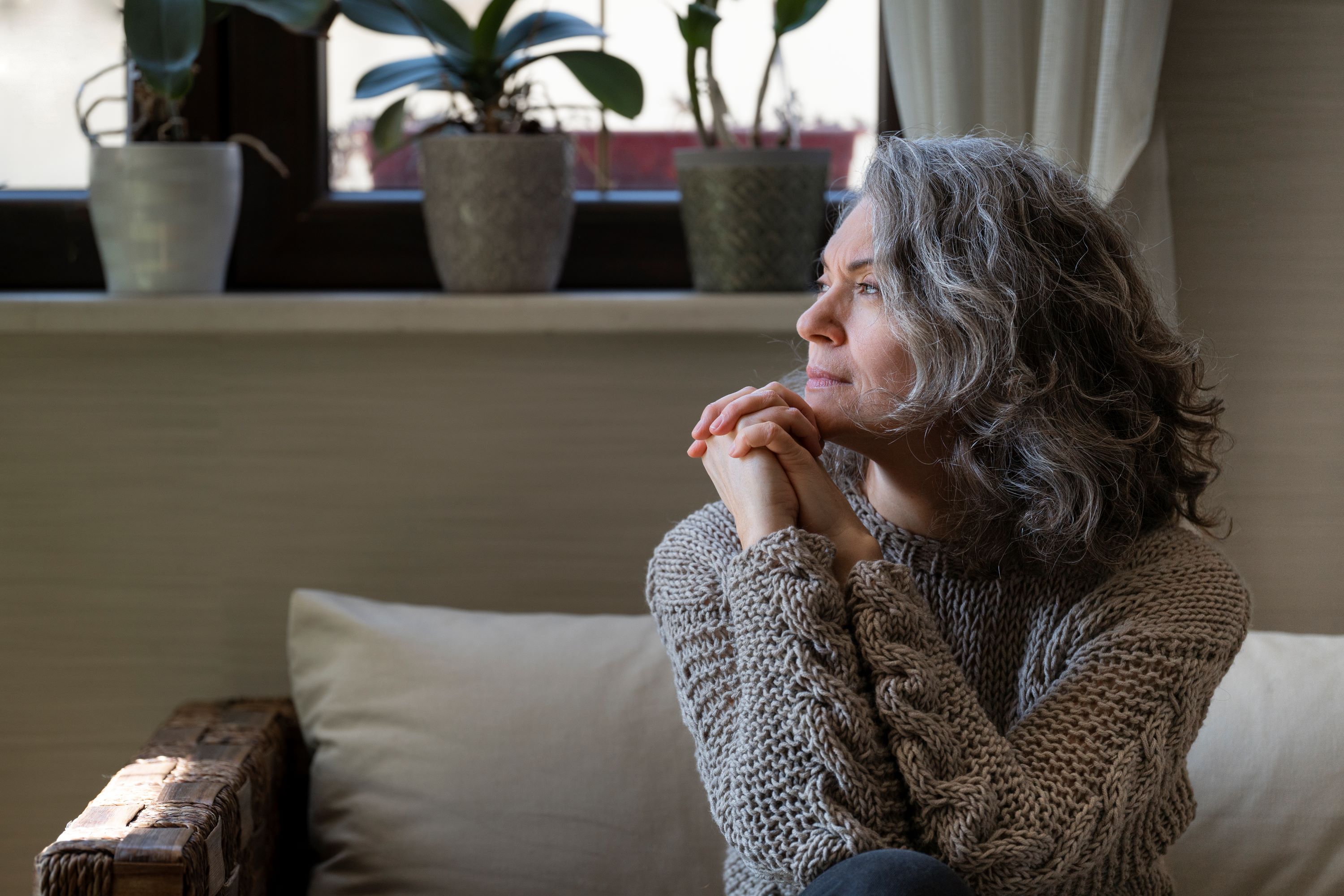 A woman with grey curly hair sits with knees up on a couch staring out to the side.