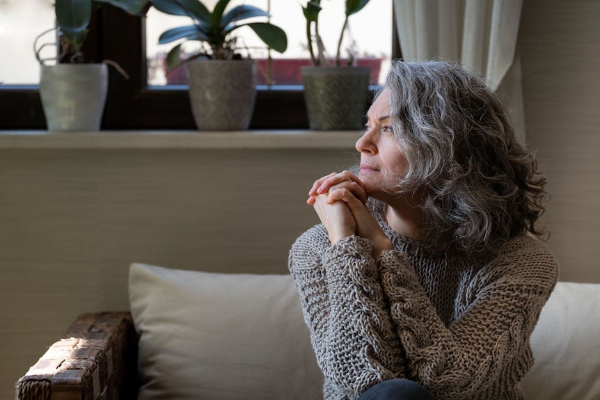 A woman with grey curly hair sits with knees up on a couch staring out to the side.
