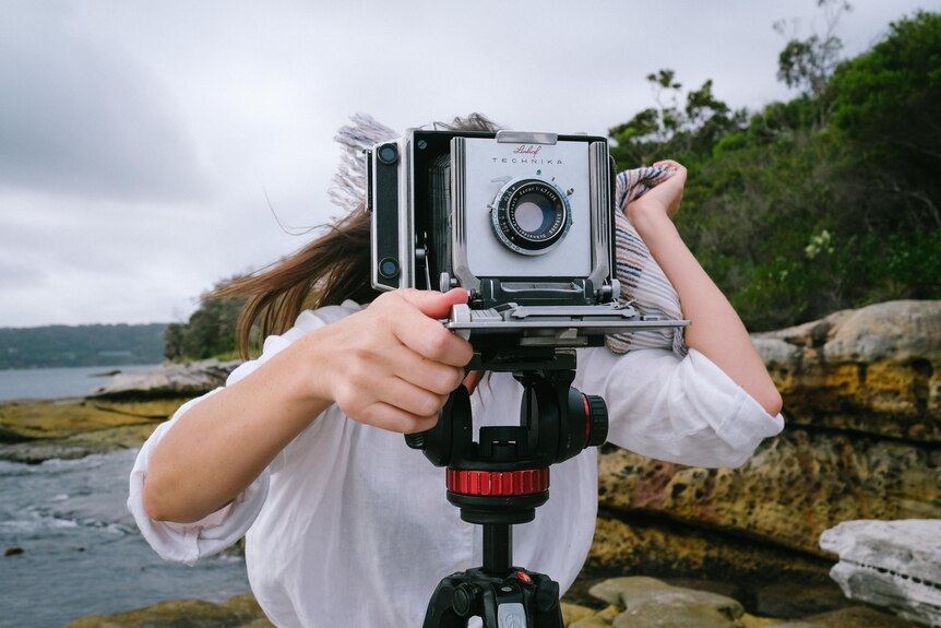 Sammy Hawker stands on the edge of Sydney Harbour balancing a large format camera on a tripod