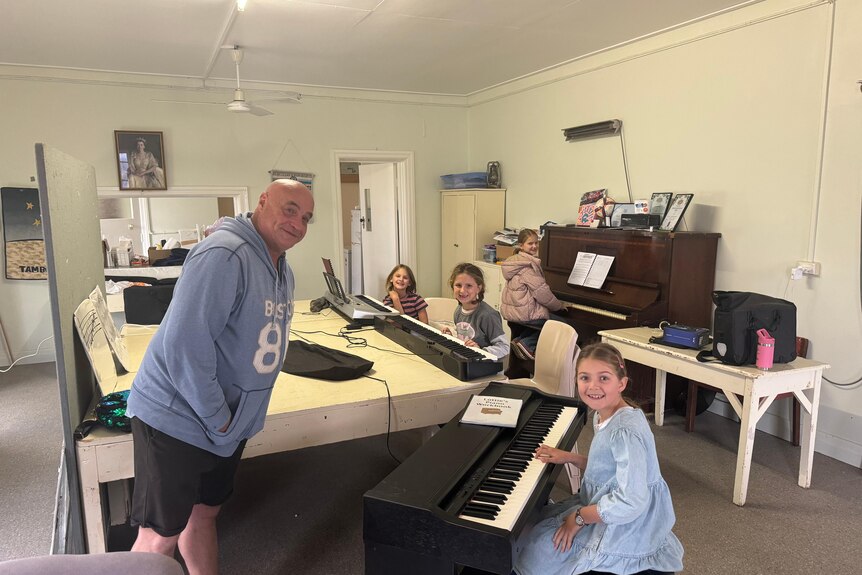 Mark McGurgan stands over four girls as he teaches them piano. They all are smiling at the camera.
